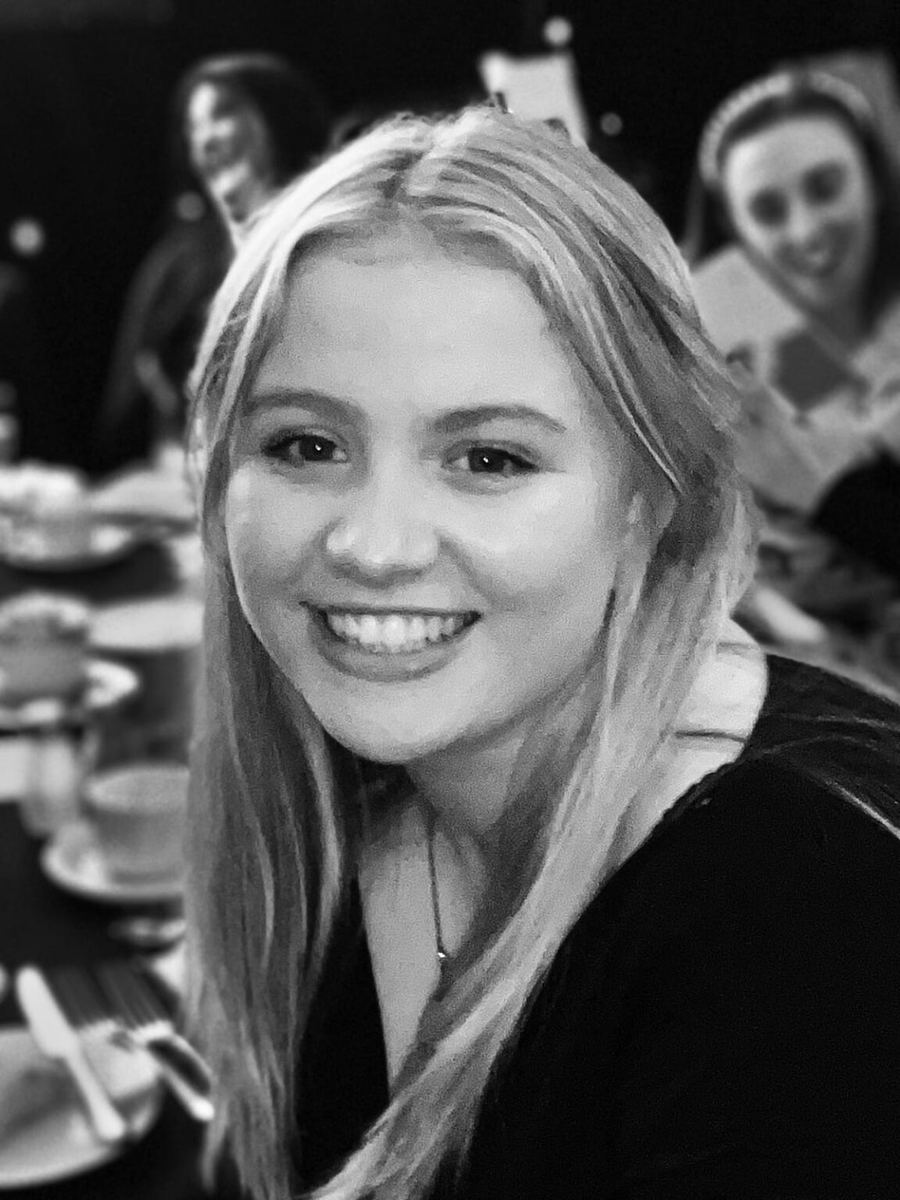 Black and white photograph of Mia, smiling towards the camera, sitting at a table laid with cups and saucers, with two people blurred in the background.